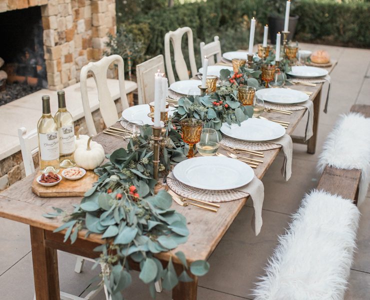 This fall tablescape is set up outside with a long, rustic wood table.One side of the table has chairs and the other side has a bench with white faux fur pelts. The tablescape has white plates, amber glass goblets and a large, leafy garland running along the length of the table.