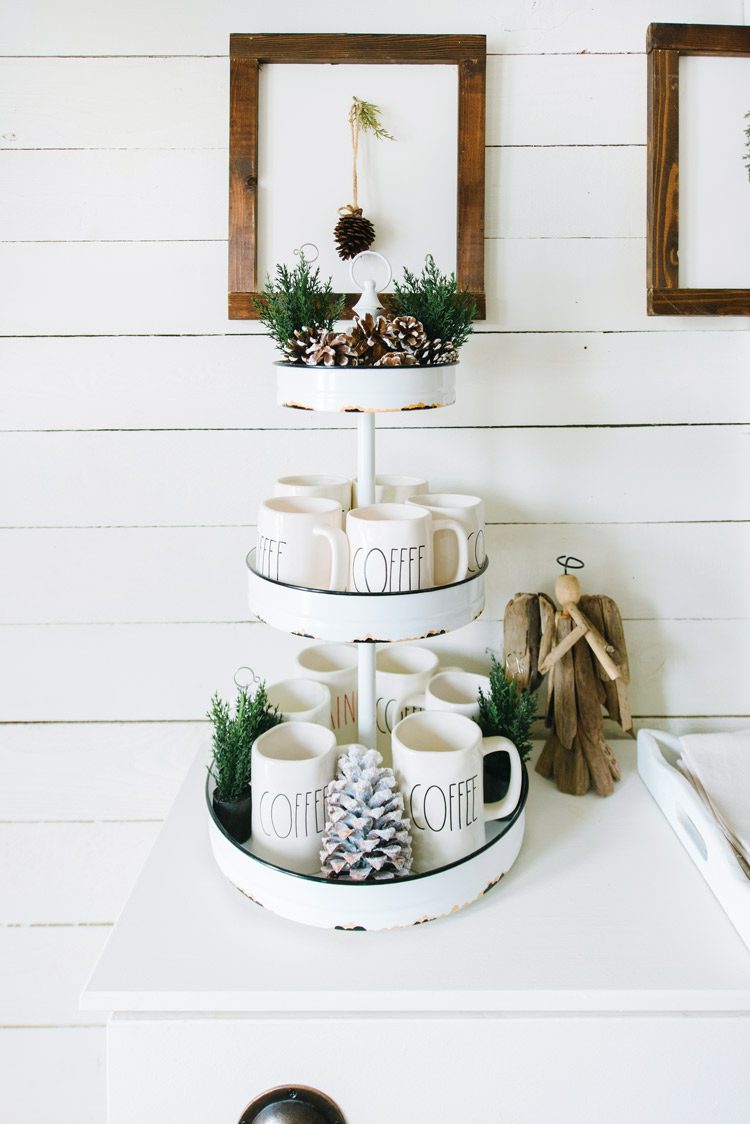 Three-tiered white tray with black edges, holding Rae Dunn mugs, pinecones and spruce tips.