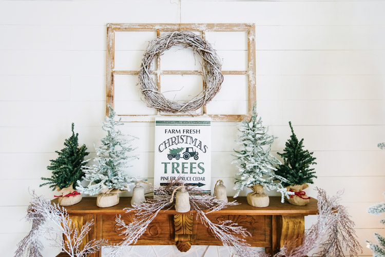 Side table decorated with mini trees and vintage-inspired decor. A wreath hangs above it.
