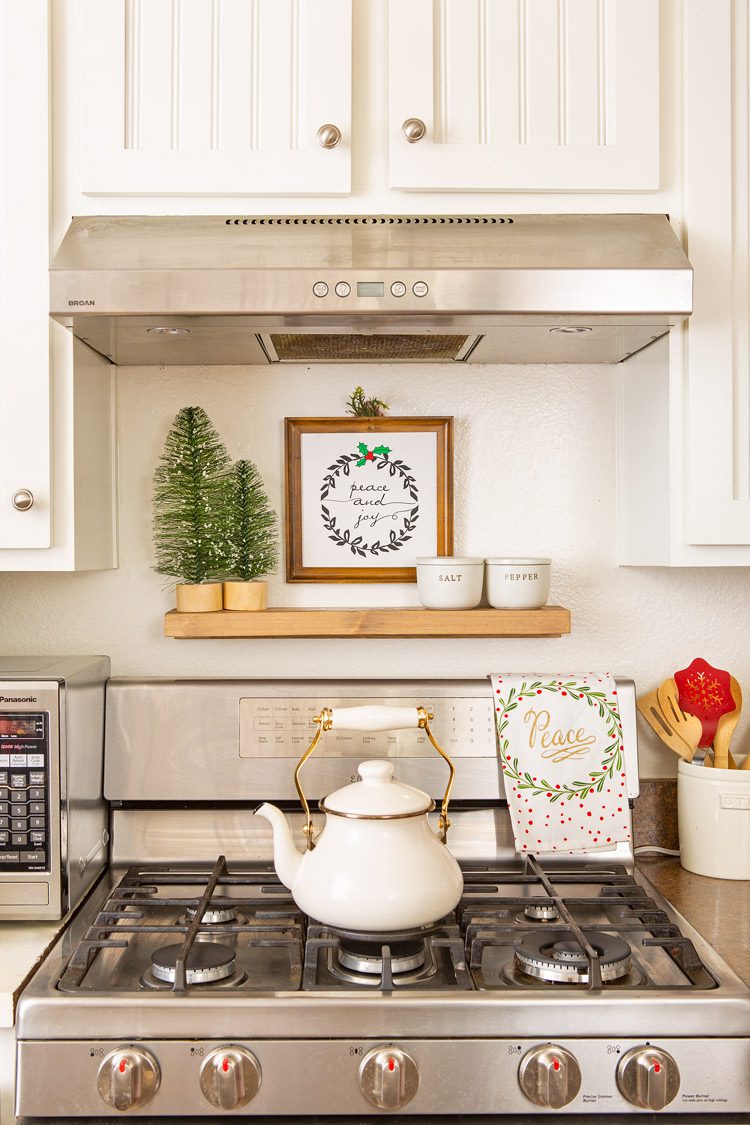 An off-white tea pot on the gas stove with a wooden Etsy holiday sign above it.