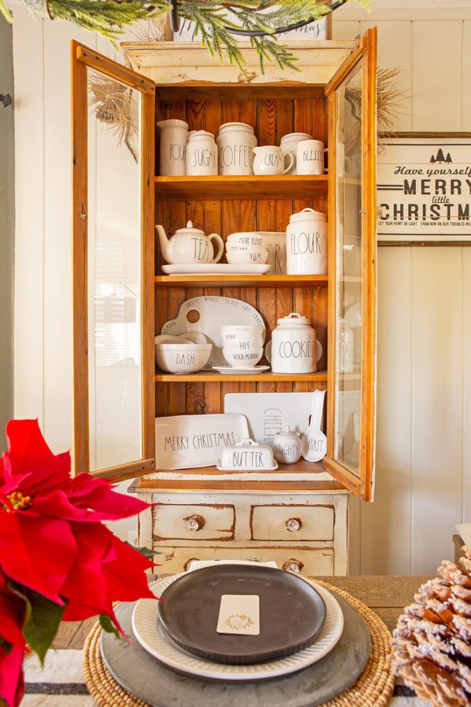 A collection of Rae Dunn ceramics in an opened wood cabinet in a Christmas cabin