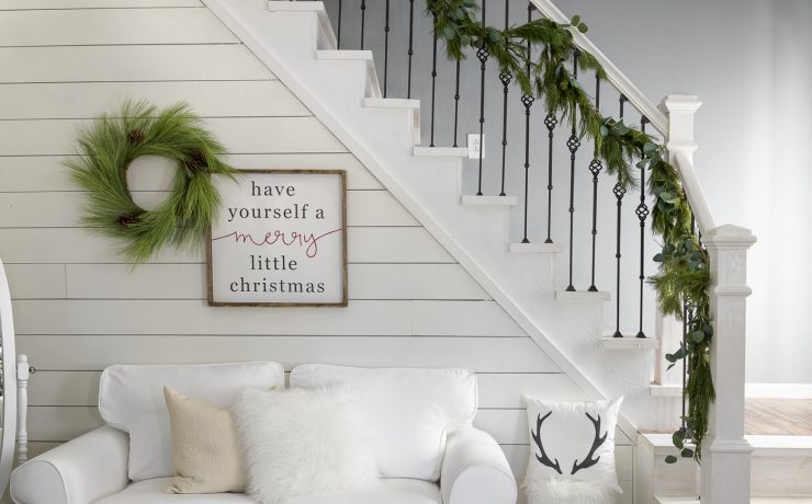 Small, white couch beneath a garland-adorned staircase and white shiplap wall, perfect for the neutral Christmas look.