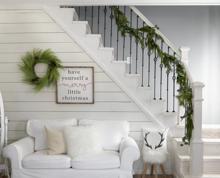 Small, white couch beneath a garland-adorned staircase and white shiplap wall, perfect for the neutral Christmas look.