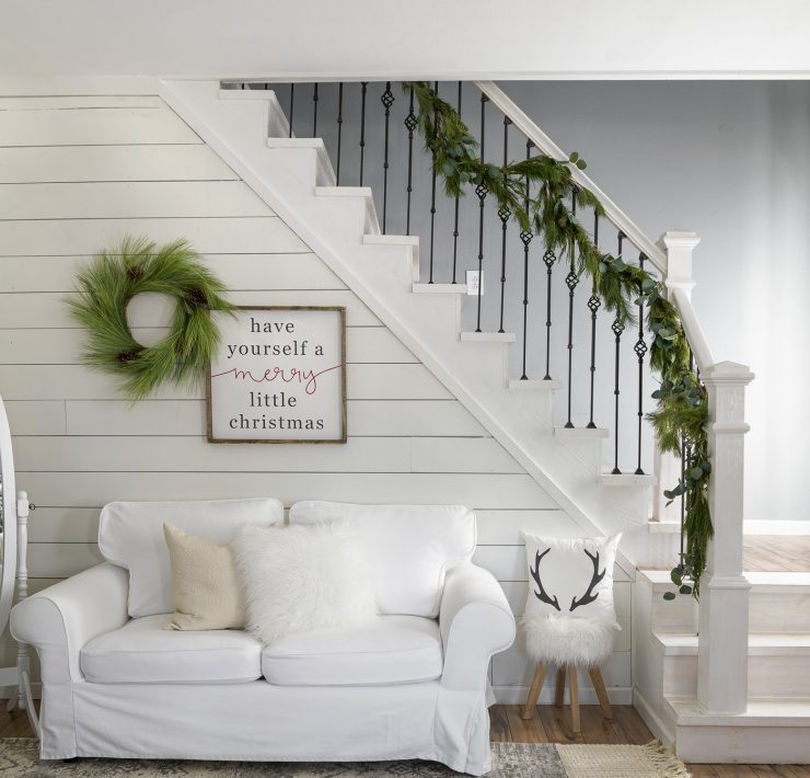 Small, white couch beneath a garland-adorned staircase and white shiplap wall, perfect for the neutral Christmas look.