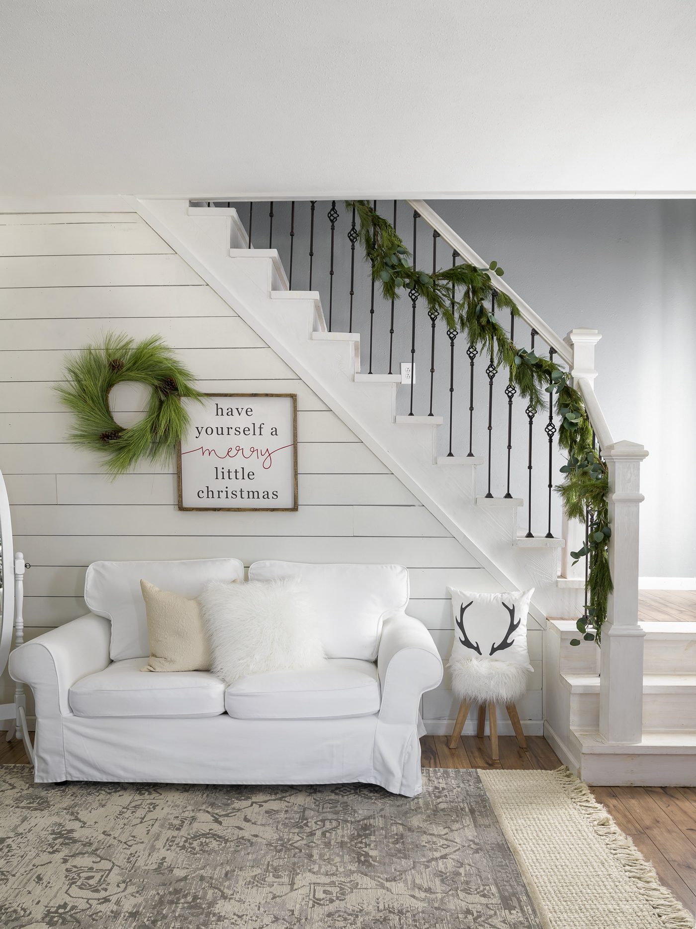 Small, white couch beneath a garland-adorned staircase and white shiplap wall, perfect for the neutral Christmas look.