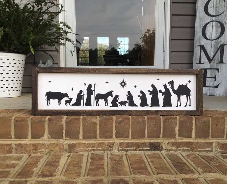 Painted black silhouettes of the nativity scene on an off white background of a dark-stained wood frame.