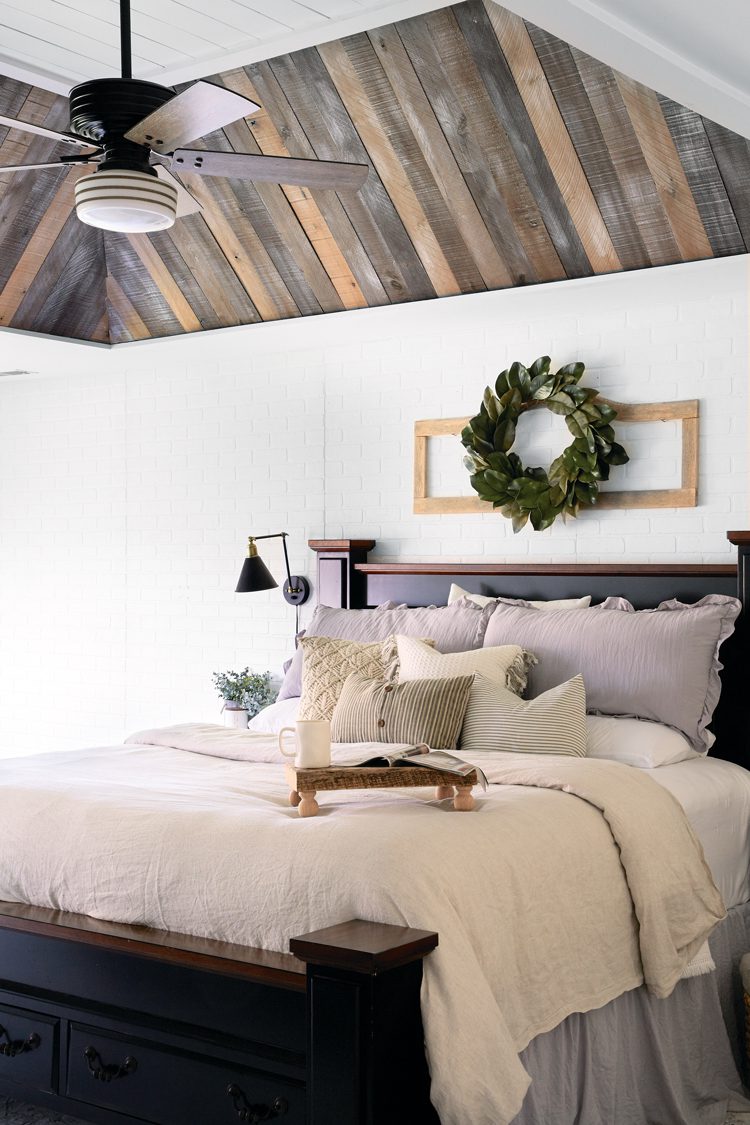 A dark black bed frame with white and light gray bed sheets and comforter. above the bed hangs a wreath made of green leaves. The room is white