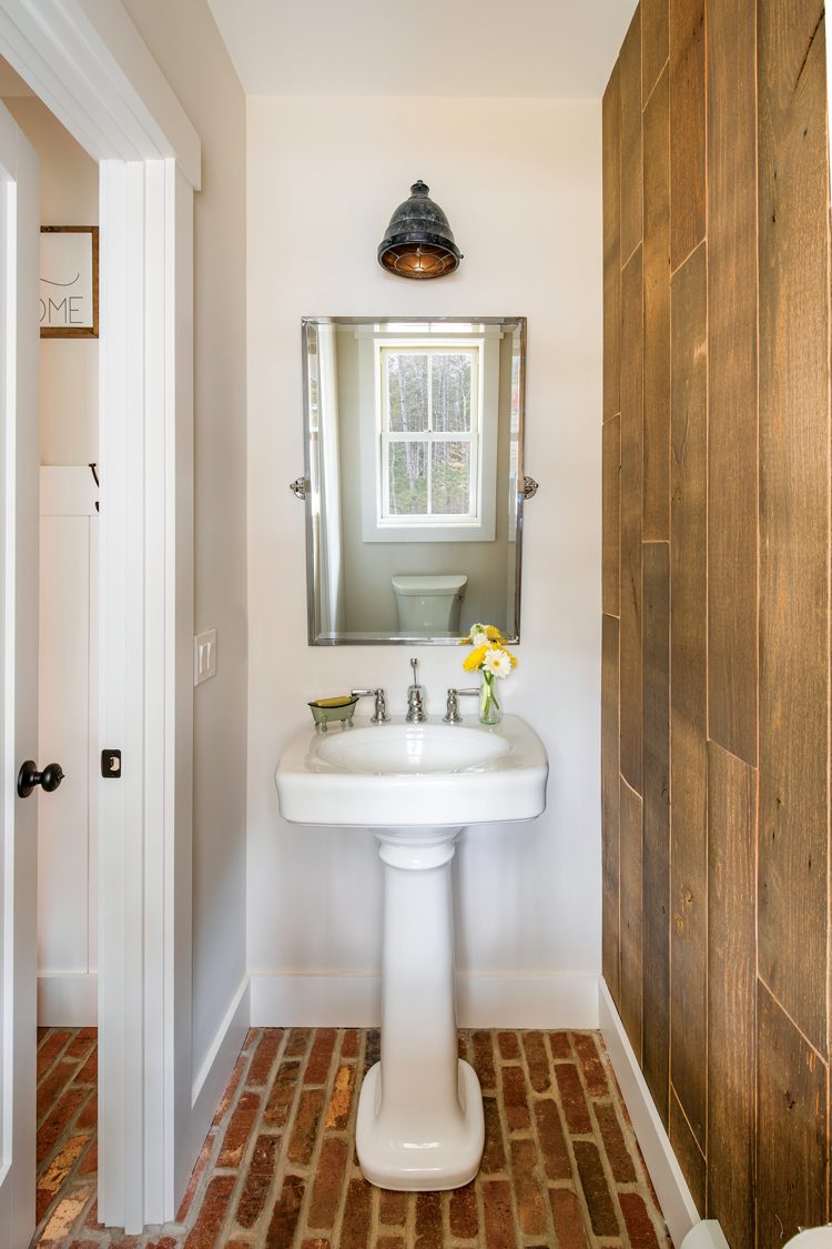 The small bathroom has exposed brick flooring with a wood accent wall. A tiny white sink sits under the small vanity mirror