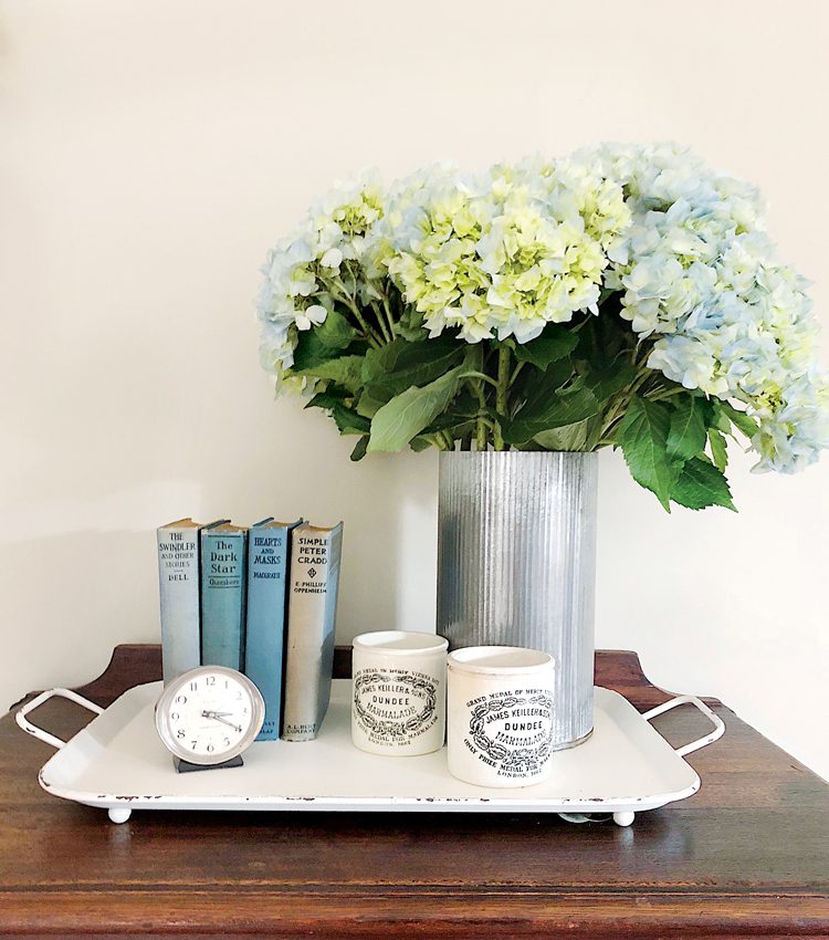 Four vintage books in varying shades of blue and gray next to a bouquet of mums and two vintage English advertising pots