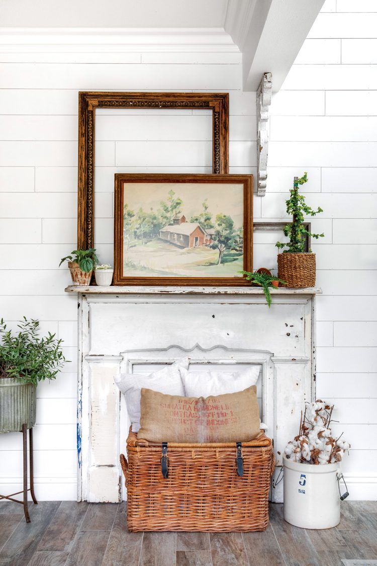 A faux white fireplace. On top of the fireplace are potted plants and a wood frame with a drawing of a classic red school building from the nineteenth century