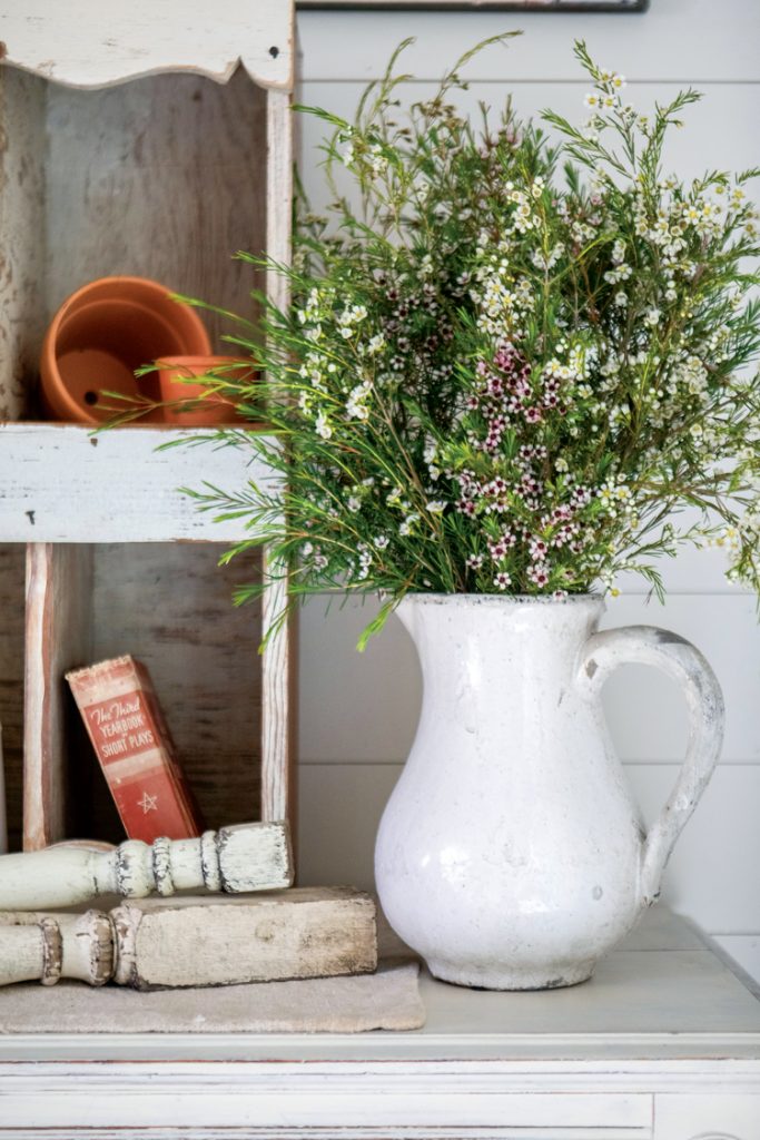 A white jug full of green petite leaves on top of an antique white shelf