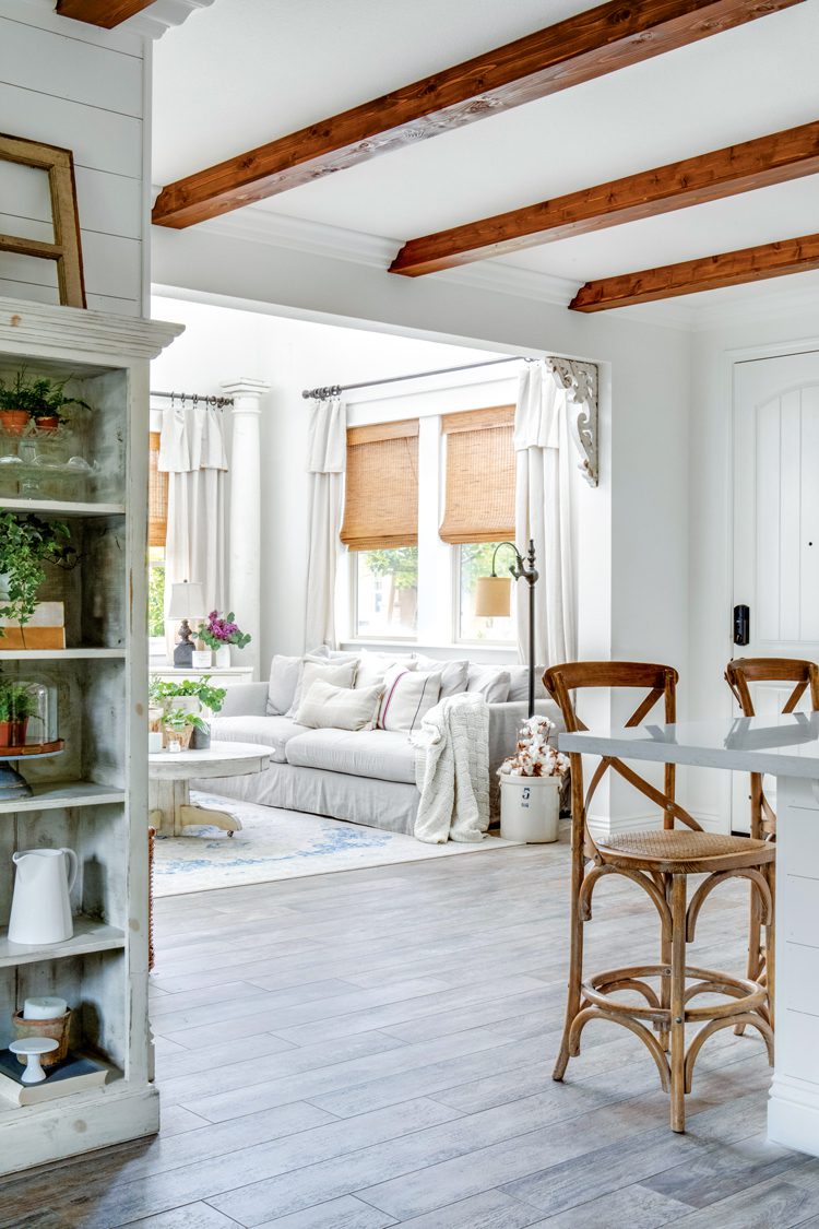 A white room with dusty gray wood floor and on the ceiling there is exposed wood beams. A white couch sits in front of open windows with natural light