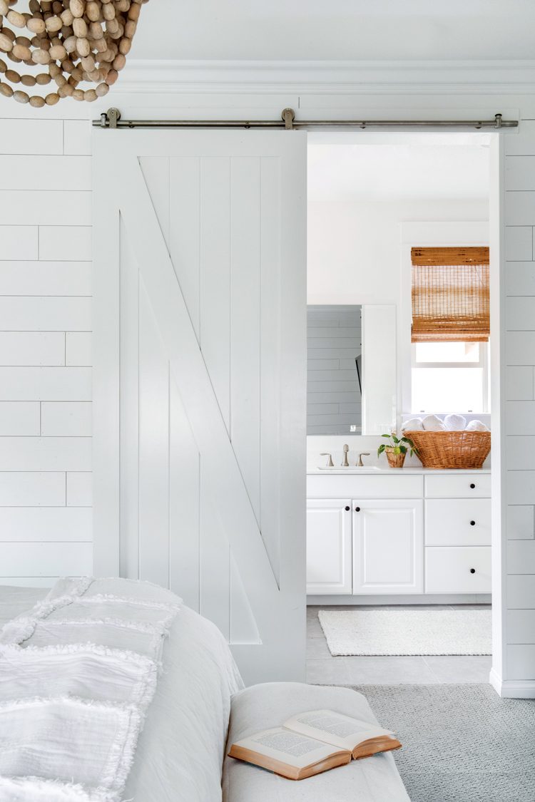 A white sliding barn door opens to reveal a white bathroom with white counters and wicker basket accessories