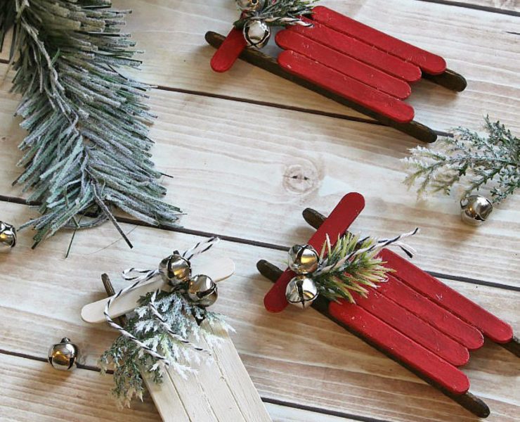Three DIY ornaments made with painted popsicle sticks, spruce tips and mini silver bells made to look like sleighs.