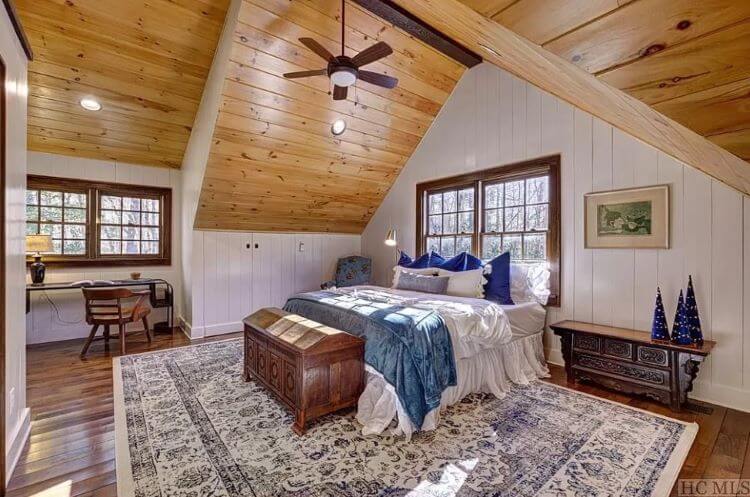 Large vaulted ceiling in the master bedroom with rich cherry oak wood and white shiplap paneling on the walls. Behind the bed is a large window with pristine views of the woods and lake
