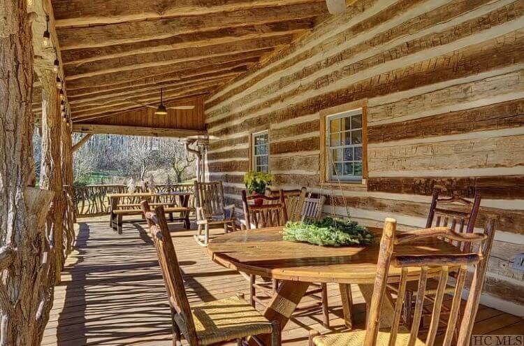 The large cabin porch with wood logs and mortar. Many rocking chairs and two tables sit on the large deck