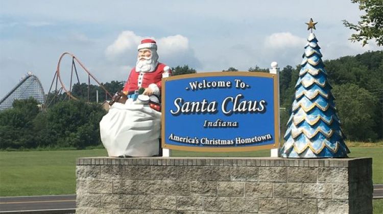 The sign to Santa Claus, Indiana, with a statue of Santa and a tree with the Holiday world rollercoasters in the background