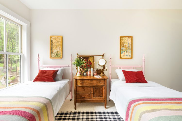 The upstairs bedroom has two twin beds. Each bed is identical with pale pink wire bedframes and a rustic wood nightstand covered in plants, mirrors and an oil painting of roses