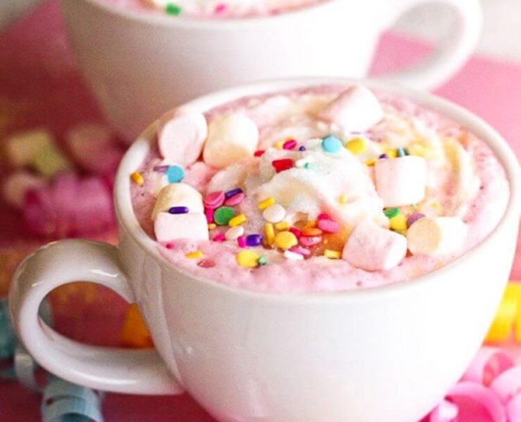 Pink foamy hot chocolate is mixed with rainbow sprinkles and mini marshmallows