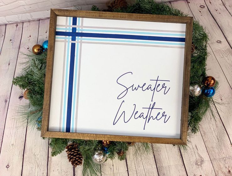 Wood-framed plaid decor piece with two intersecting blue plaid stripes and "sweater weather" in calligraphy.