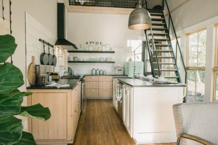 A modern farmhouse kitchen with dark countertops and a blue fridge next to a stairs that operate on a pulley system for the sake of space in this tiny home