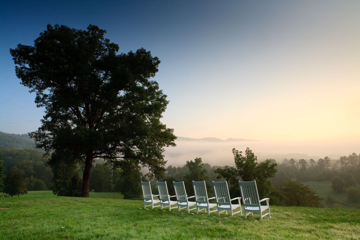 A view of rollign green hilsl from a veranda with a row of white farmhouse style lawn chairs