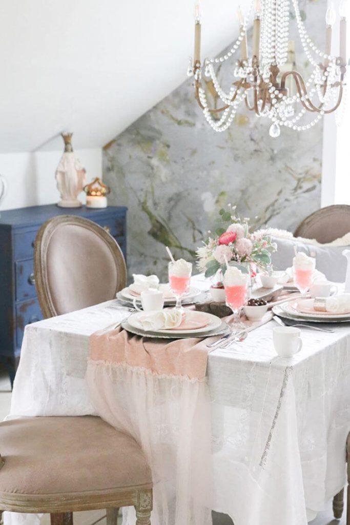 A sparkling glass chandelier hangs above a farmhouse table decked out in a pink velvet table runner and silk pink table settings. There is a bare wood centerpiece in the middle of the table under a bouquet of flowers.