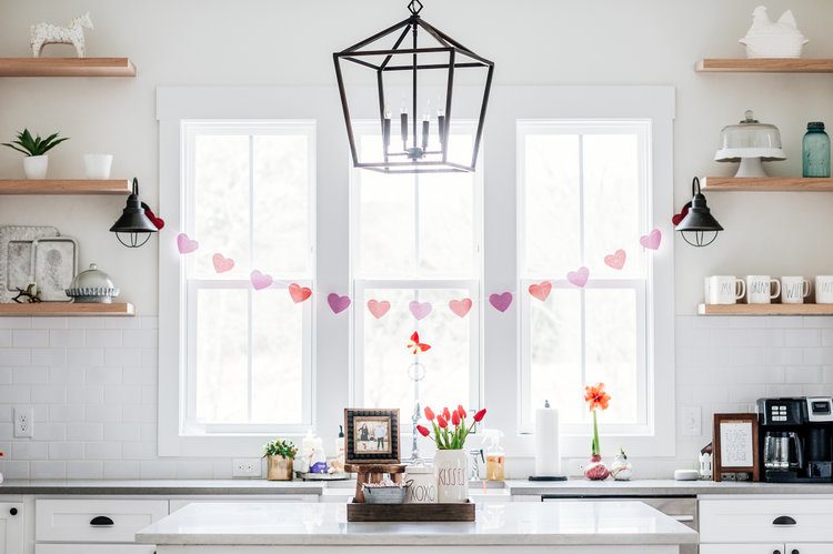 Bare farmhouse kitchen shelves are connected by a strand of hearts on a garland in shades of hot pink and reds. Bare blooms are used in vases on the kitchen island.