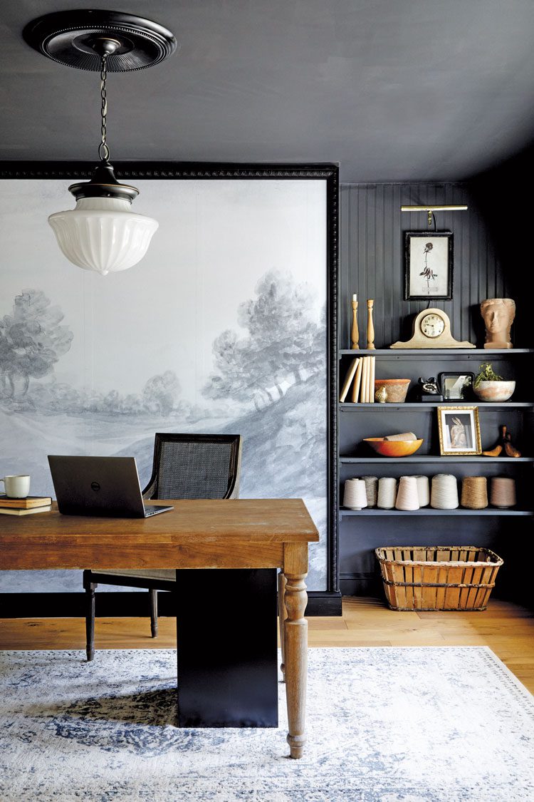 A faded rug rests under the Old World inspired wood table. An oversized painting rests on the floor behind the table and reaches the ceiling. The picture is of a gorgeous grey and white provincial scene in rolling hills 