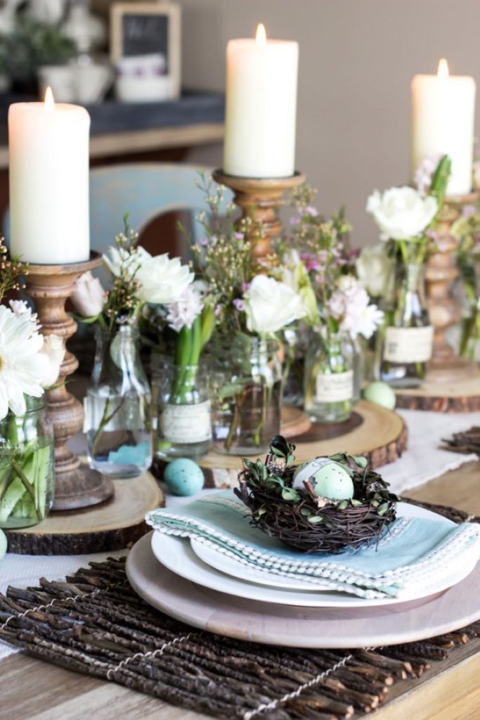 A rustic tablestable with tall votive candle stands in wood holding white candles. Each table settign on this tablescape has a mini nest with a faux robin egg inside.