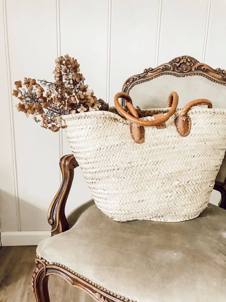 Dried florals in a woven basket with a large handle on top of an old fashioned, French style chair