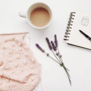 A baby pink scarf being knit with a knittign needle and a cup of coffee beside a sprig of lavender