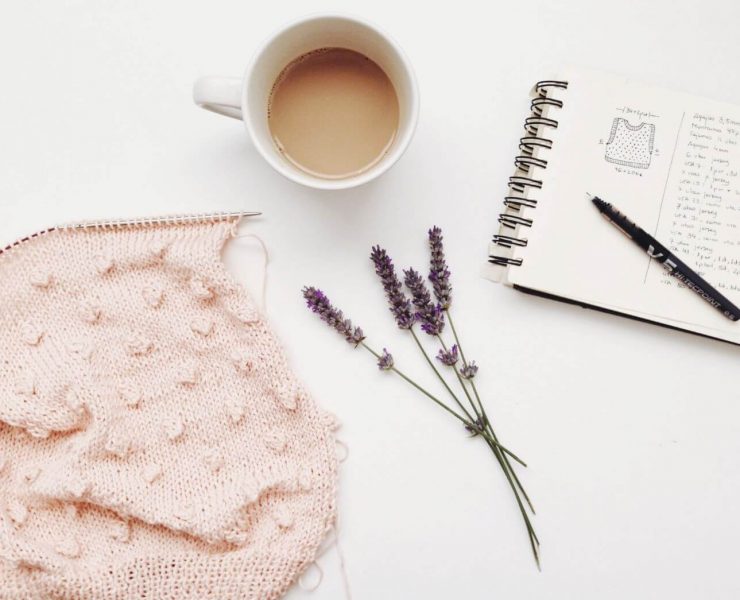 A baby pink scarf being knit with a knittign needle and a cup of coffee beside a sprig of lavender