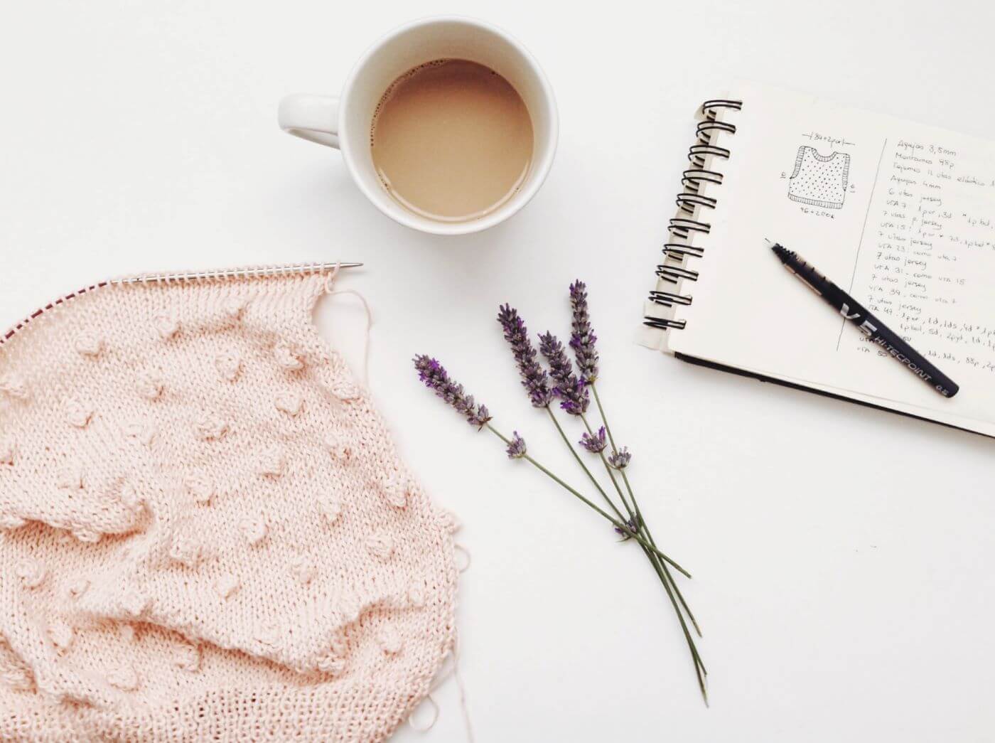 A baby pink scarf being knit with a knittign needle and a cup of coffee beside a sprig of lavender