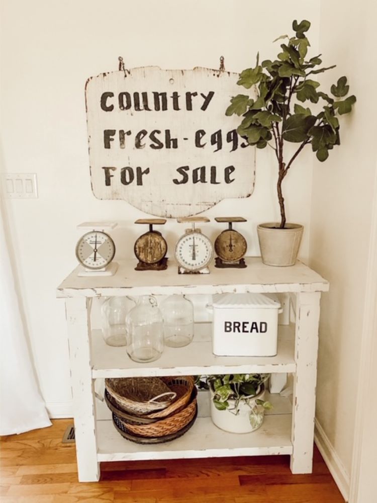 A small book shelf houses a sign that reads, “Country Fresh Eggs For Sale.” Accompanying the sign are layers of baskets and scales, originally used in markets.