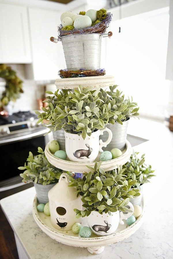 A three-teired tray has living succulant plants in mugs coverde in easter bunnies. Scattered robin eggs in many shades of blues and greens add to the display