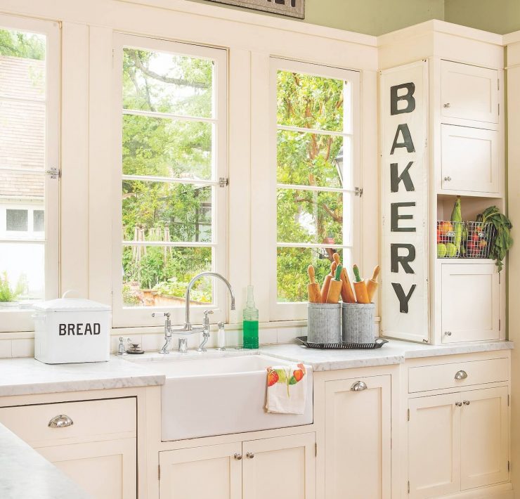 A kitchen painted in sage green on the walls and white cabinets and clean countertops. a large sign says Bakery near the clear windows.