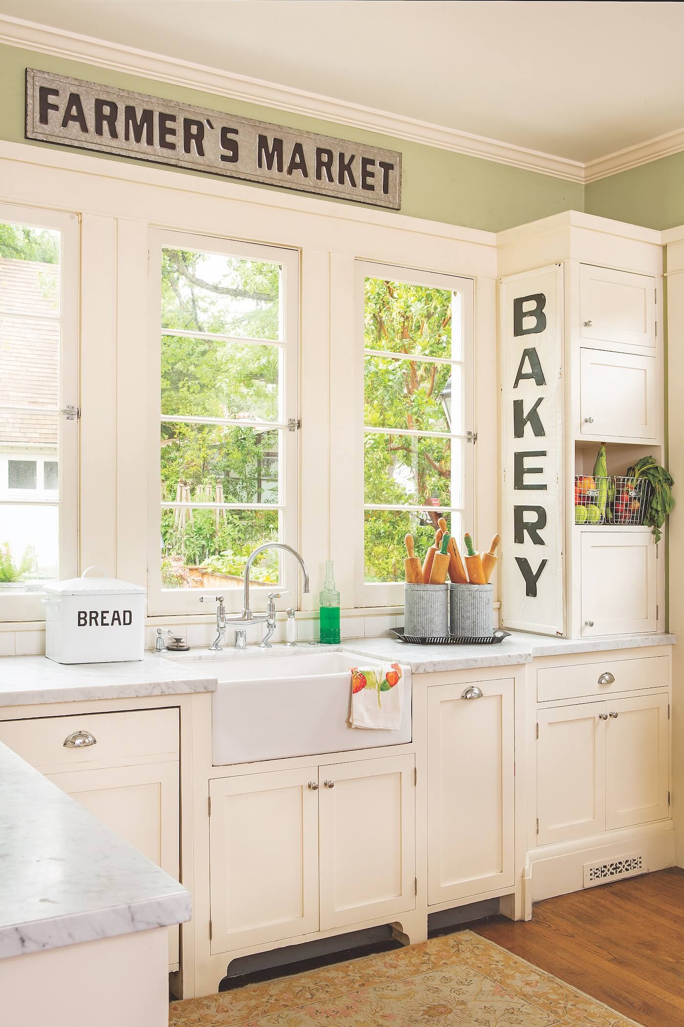 A kitchen painted in sage green on the walls and white cabinets and clean countertops. a large sign says Bakery near the clear windows.