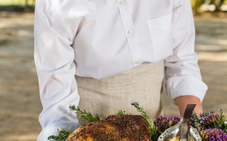 A woman is holding a platter covered in the slow roasted leg of lamb framed by freshly cut spring roses in the colors of purple and light orange