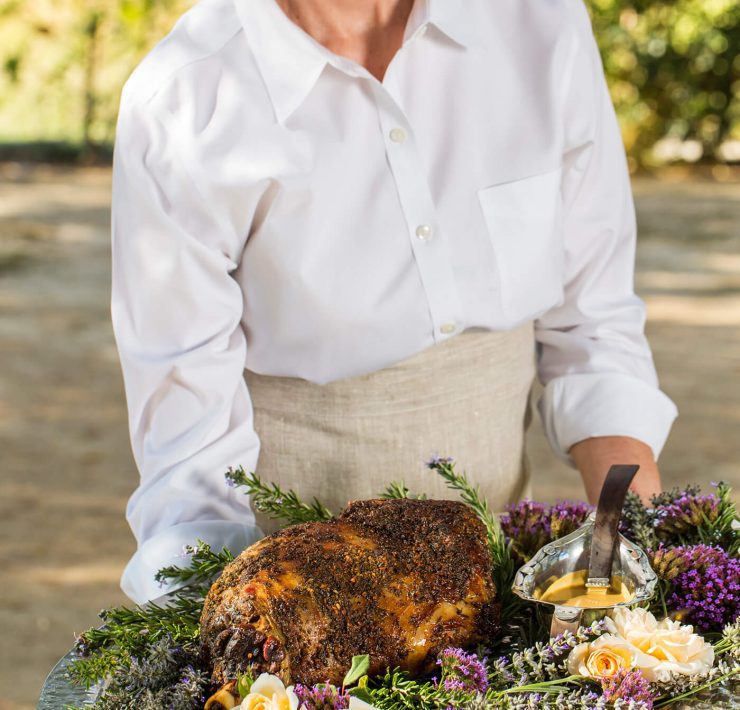 A woman is holding a platter covered in the slow roasted leg of lamb framed by freshly cut spring roses in the colors of purple and light orange