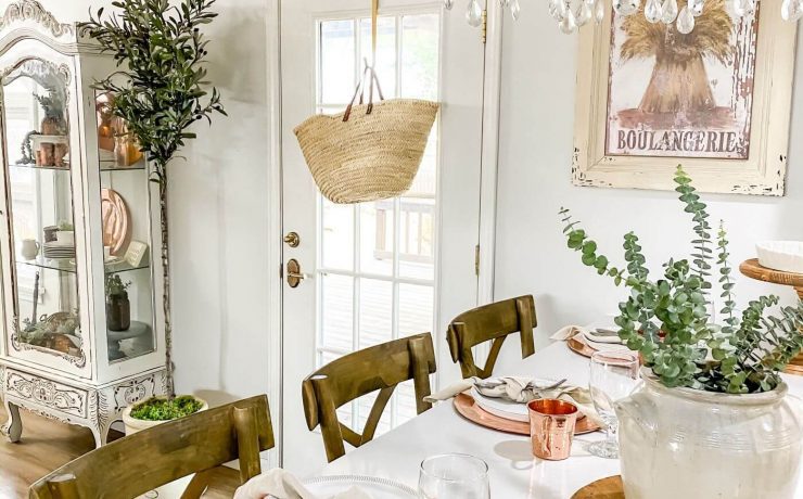 A white farmhouse table with an antique crystal chandelier above. The table settings are made from copper colored material and a white ceramic piece houses fresh pops of green from the garden in the table’s centerpiece