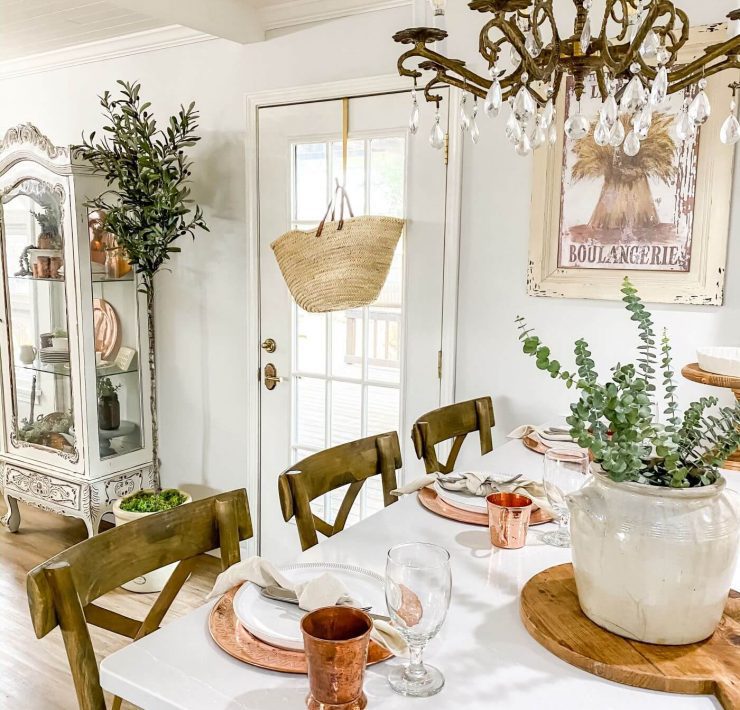 A white farmhouse table with an antique crystal chandelier above. The table settings are made from copper colored material and a white ceramic piece houses fresh pops of green from the garden in the table’s centerpiece