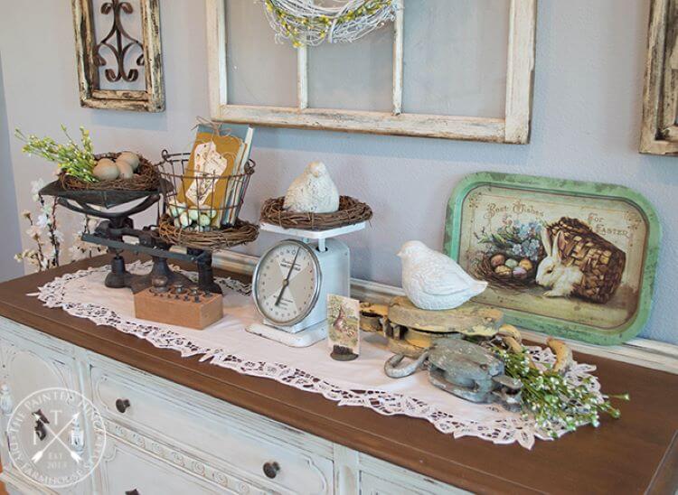 a chest of drawers is adorned in a lace-lined runner covered in vintage pieces, like three antique scales each housing a nest with a stone bird inside for Easter decor