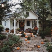 A white rustic style fall she shed with a porch is surrounded by mums and pumpkins