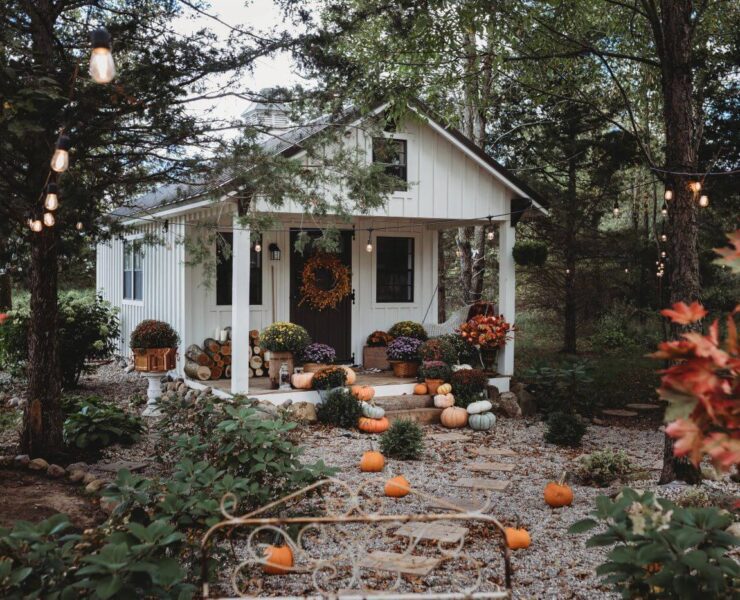 A white rustic style fall she shed with a porch is surrounded by mums and pumpkins