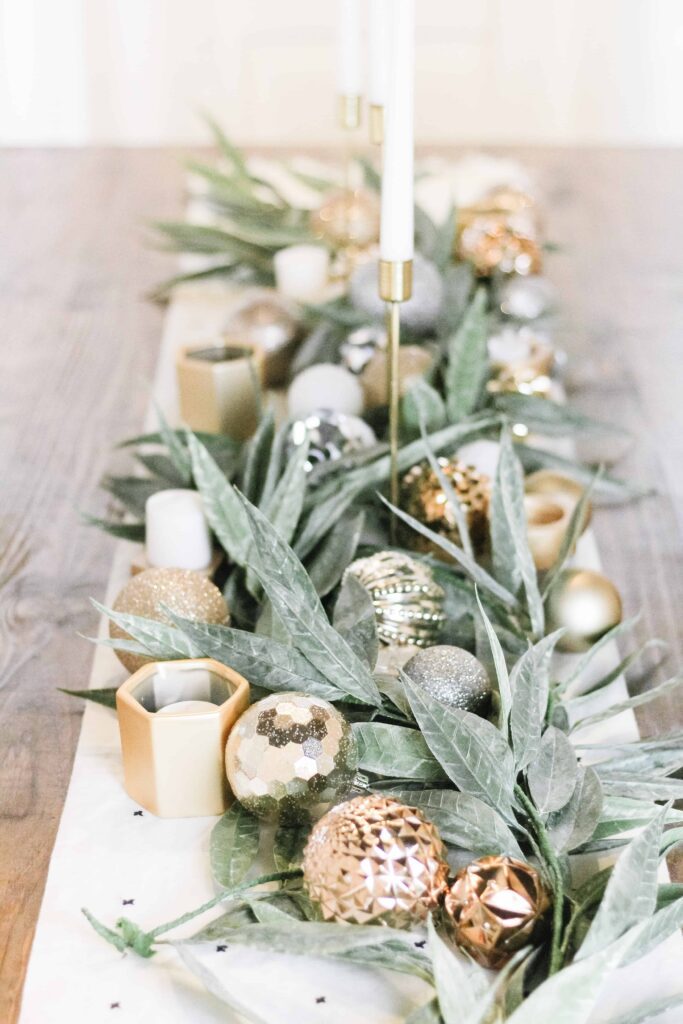 close up of center tablescape full of fresh green garland, gold ornaments, and simple white candles with a white table runner