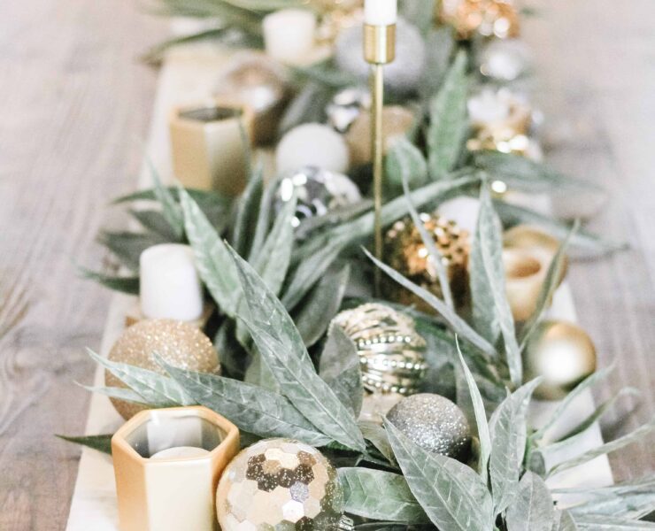 close up of center tablescape full of fresh green garland, gold ornaments, and simple white candles with a white table runner