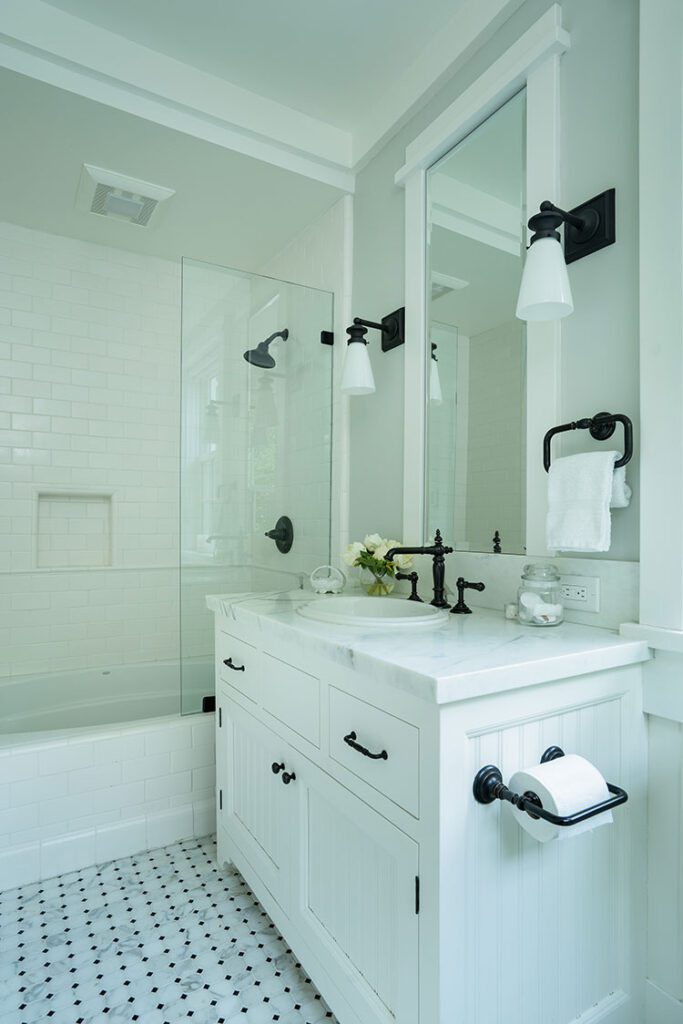 The mostly all white bathroom has pops of black in its choice of hardware and scones. Small black pieces of tile mix with white to offer more pops of dark color on the floor.