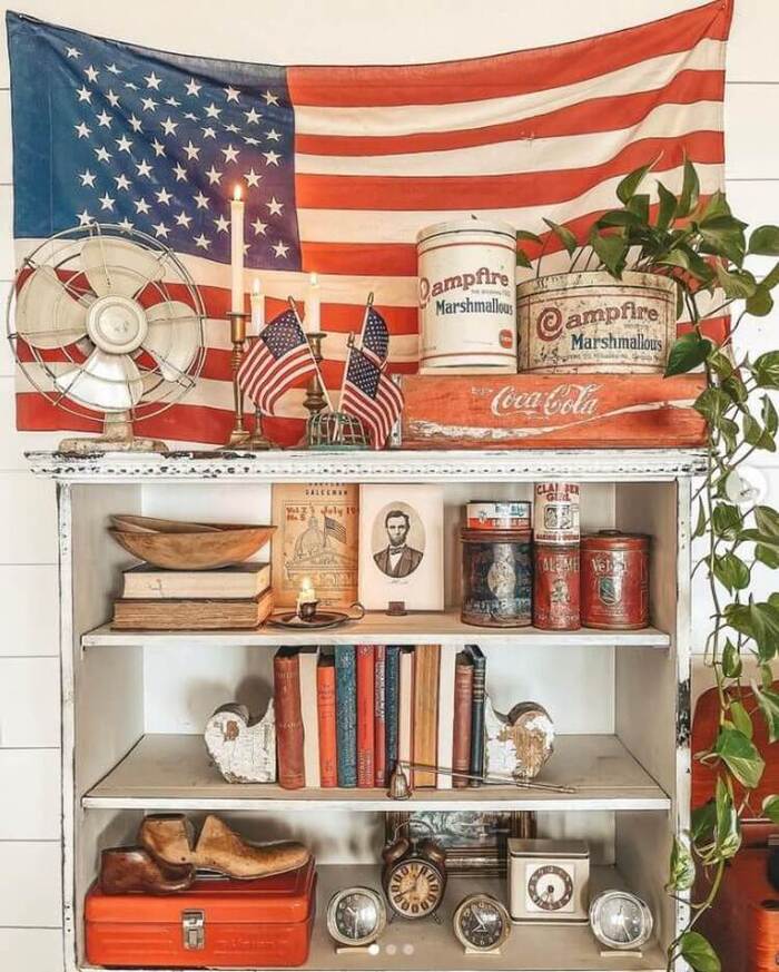 Distressed white bookshelf with 4th of July decor, like antique red and white tins, books, and other trinkets.