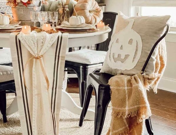 Muted colors adorn the table with dried flowers in a centerpiece and pale green and orange pumpkins. Black chairs circle the white table draped in burlap like runners.